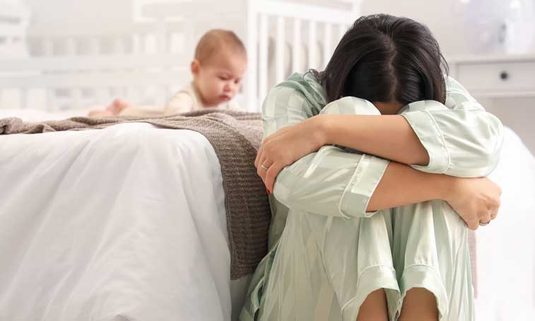a young mother experiencing mental health difficulties expressed with a young woman sitting on the floor with her head down next an infant crawling on the family bed beside her