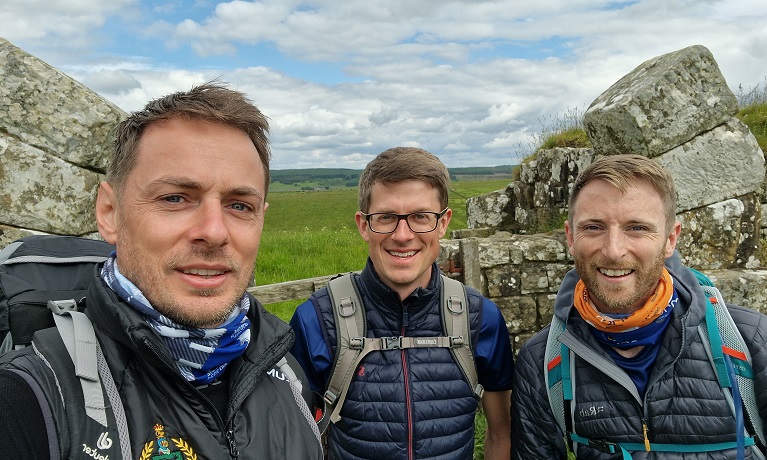 Three men wearing walking gear standing in front of a wall in the countryside
