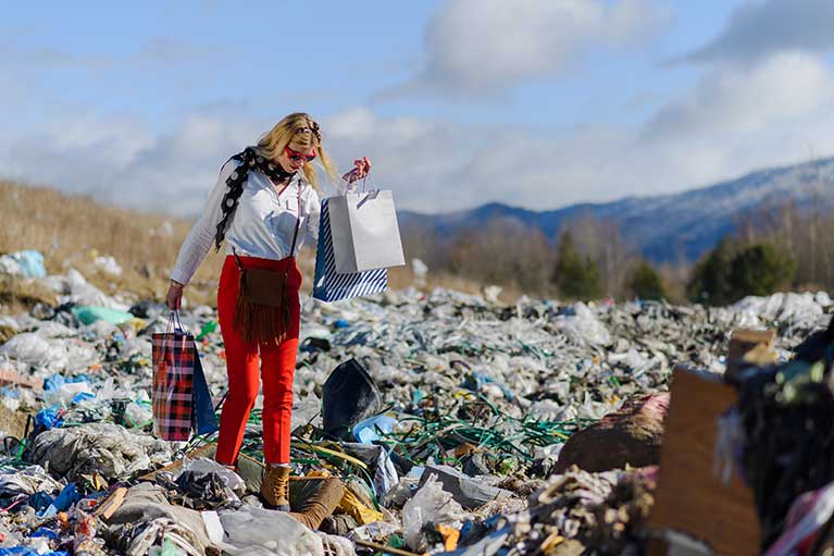 fashionably dressed woman with sunglasses carrying fashion brand shopping bags walking across a massive textile dumping ground