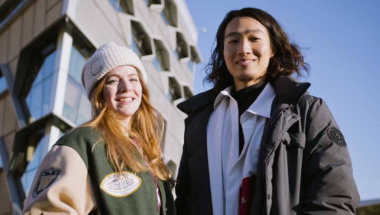 A couple of students looking down at the camera facing up with a tall building in the background