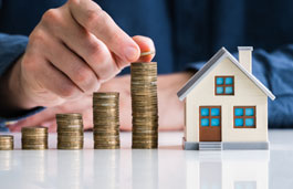 Five stacks of coins with a hand reaching for the highest stack, next to a model house same height as tallest stack