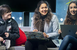 Three students sitting in front of a big screen tv with macbooks