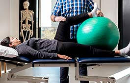 Patient lying on a physio bed with a practitioner moving patient's leg on top of a large green ball 