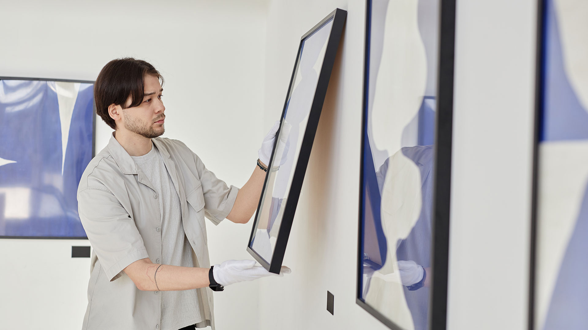 A man wearing white gloves adjusts the hanging of a painting in a gallery