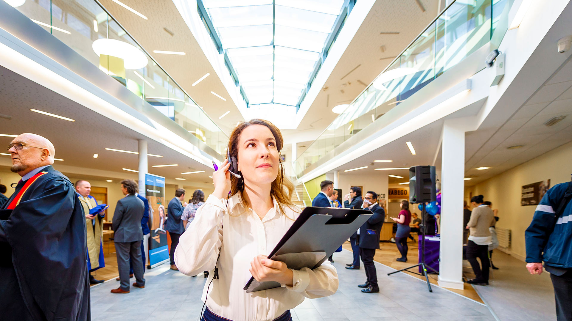 Female student standing in a busy room holding a clipboard and talking on a headset