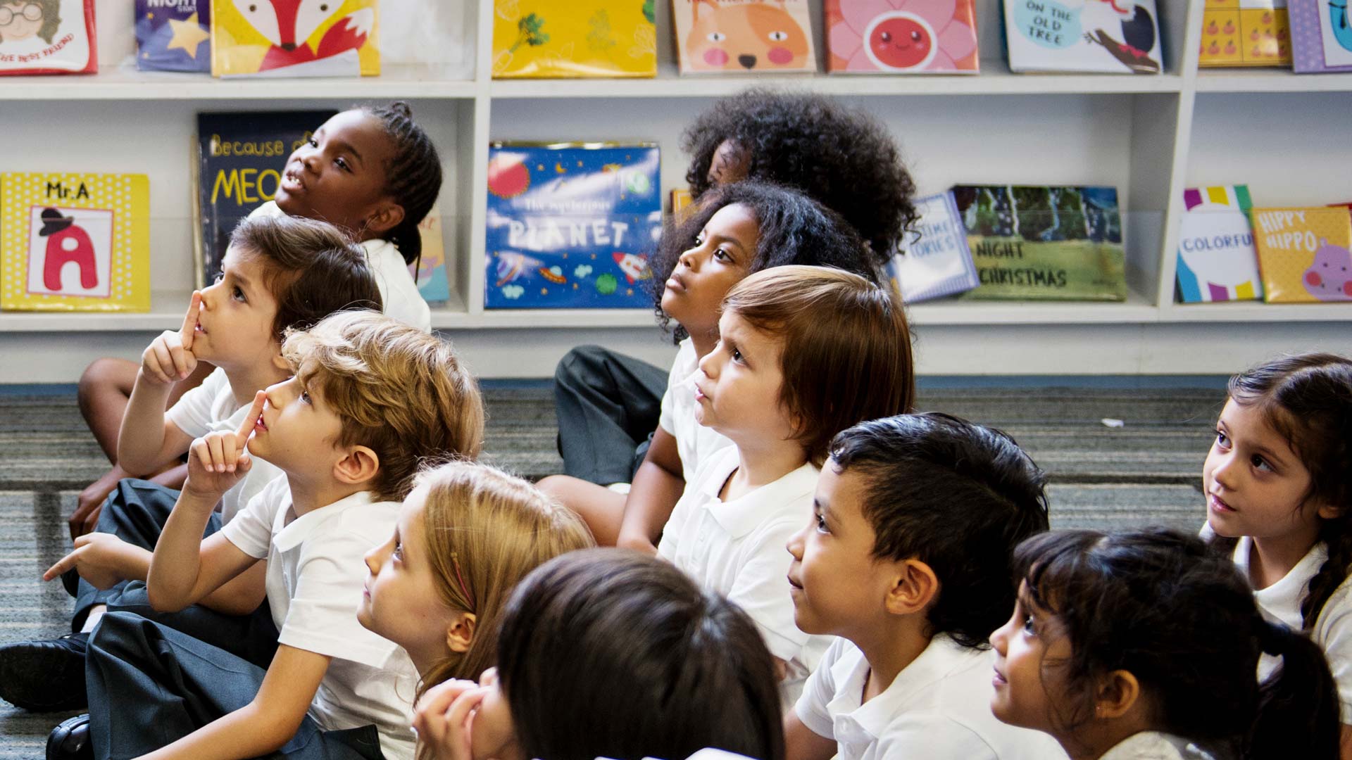A teacher at the front of a classroom talking to a group of primary school children sat on the floor.