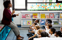 A teacher at the front of a classroom talking to a group of primary school children sat on the floor.