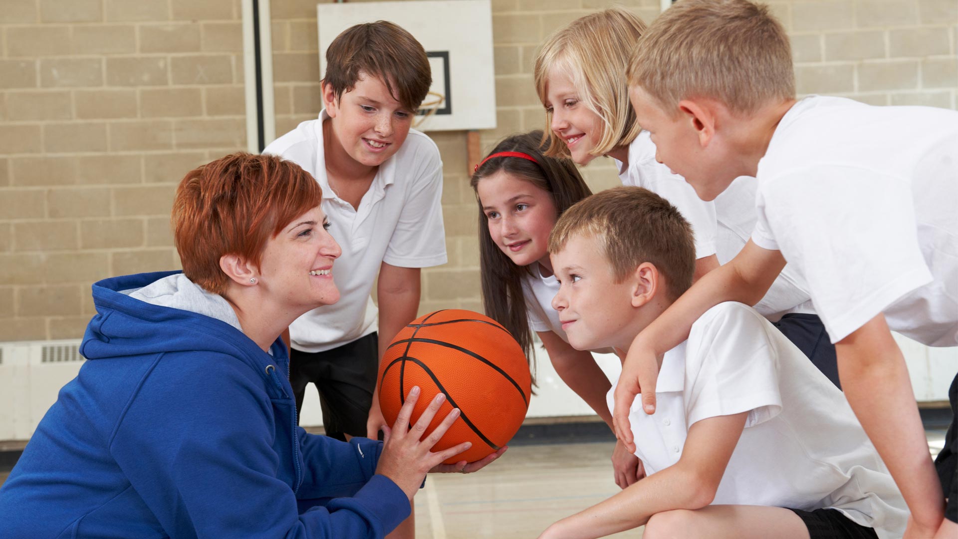 A teacher holding a basketball in a school sports hall, giving a team talk to a group of primary school-aged children.