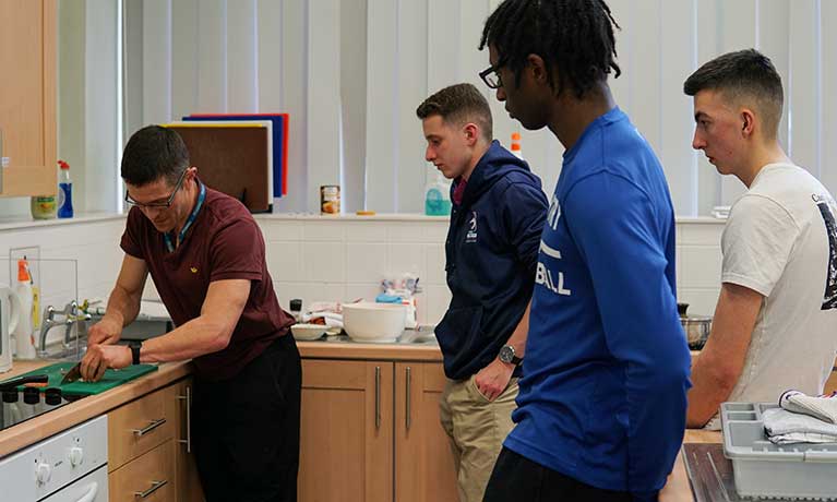 lecturer in a kitchen chopping vegetables with 3 students observing 