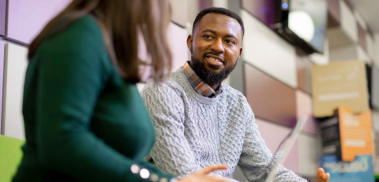 smiling male student chatting to another person with a laptop on their lap