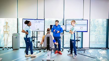 A group of three stand round a seated fourth person, discussing results shown on screens behind them