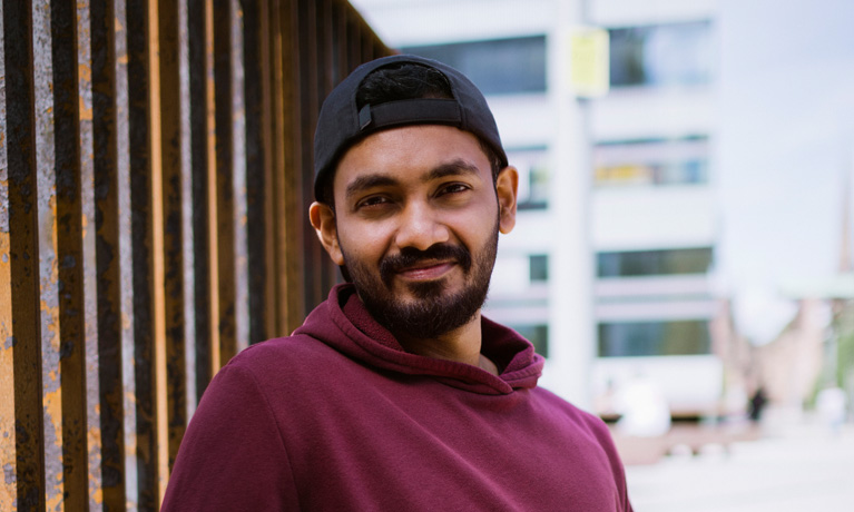 A student in a cap and maroon sweatshirt leaning against a fence.