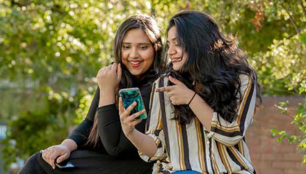 Two students smiling outside looking at a smartphone with greenery behind them.