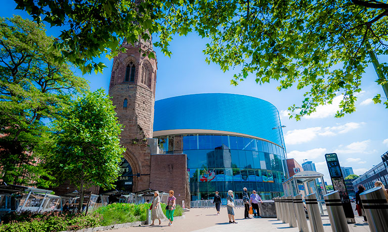 The Wave sports centre and Spire Bar in front of a backdrop of blue sky and surrounded by trees and people walking.