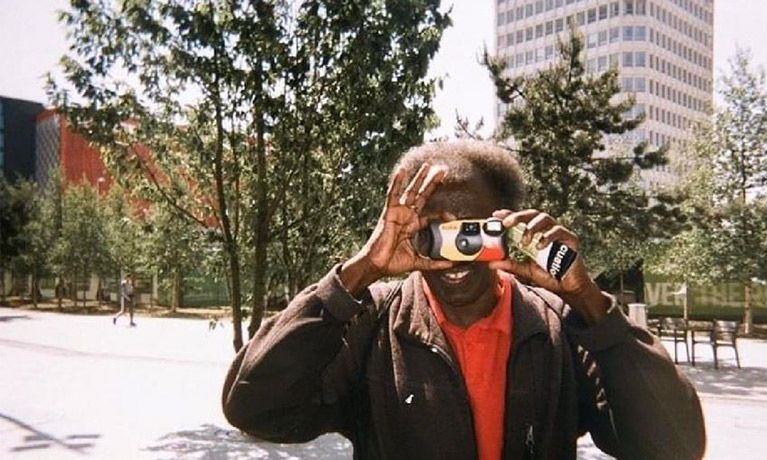 A man points  camera at the viewer in an urban landscape.