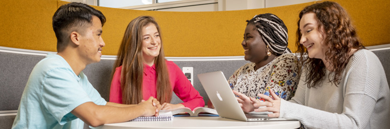 Group of four students sitting around a table chatting across their books and laptops