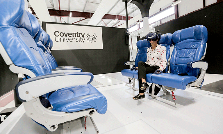 A woman sits on blue airplane style seats wearing a black VR headset