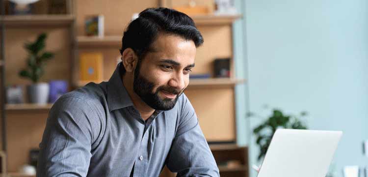 Male with dark hair and a beard working at a table in front of a laptop