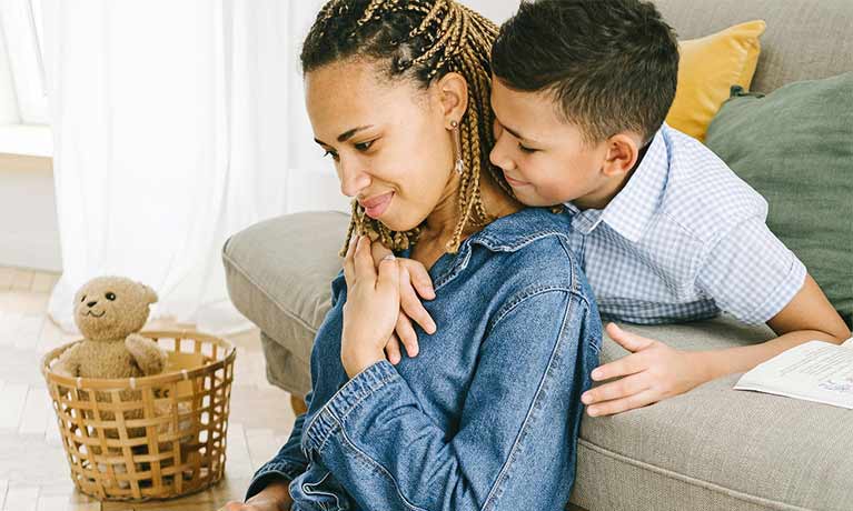 Female looking at a laptop on her lap her son is hugging her neck 