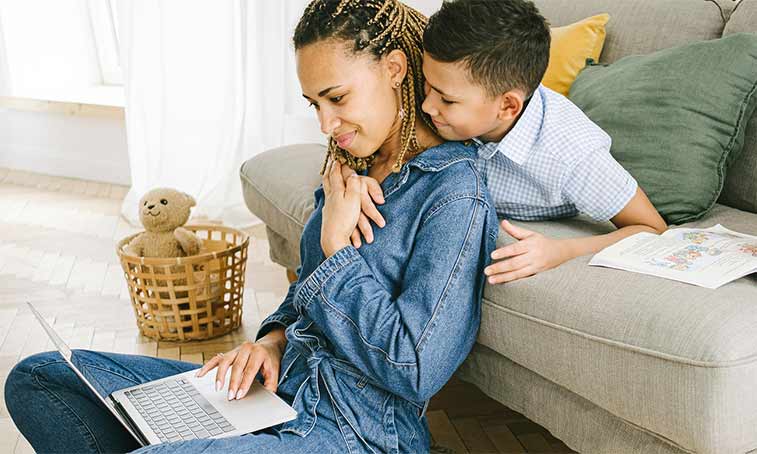 mum working on a laptop with young son hugging her neck
