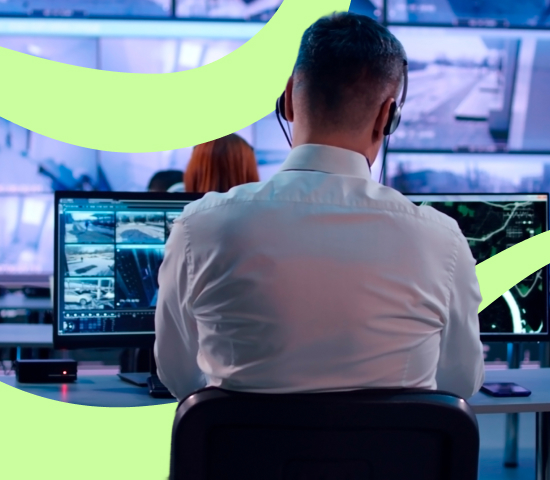 Person wearing headphones and a white shirt working at a desk with multiple monitors showing surveillance footage and data, with another person in the background.