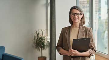 A woman with glasses holding a clip board smiling at the camera