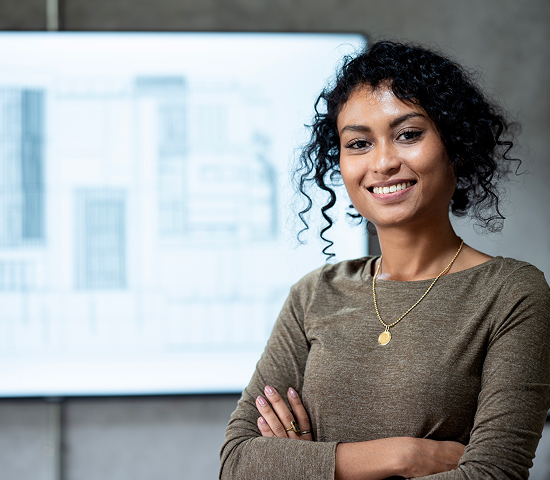 Person with curly hair and a circular pendant standing with arms crossed in front of a blurred architectural blueprint on a screen.