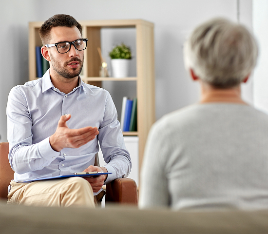 Two people talking indoors; one holds a clipboard, the other is seen from behind with gray hair.