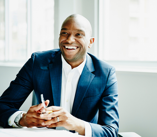 Person in a dark blue suit sitting at a table, holding a pen with hands clasped, in front of large windows.
