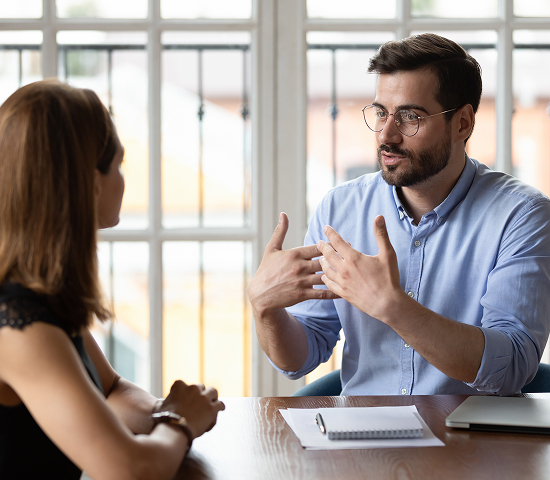 Two people talking at a table with a notebook and pen, near large windows