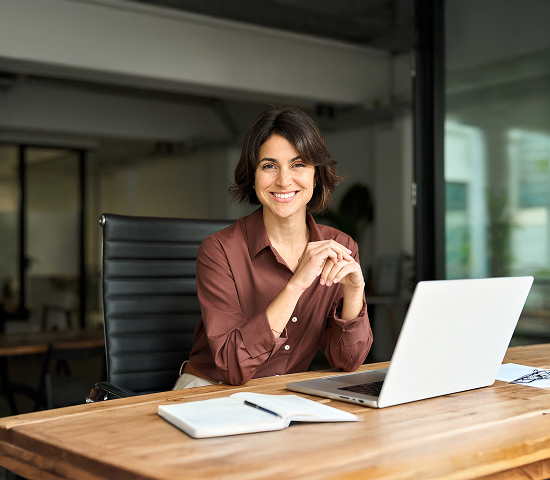 Person sitting at a wooden desk with a laptop, notebook, and papers in a modern office.