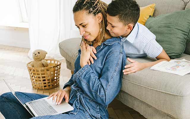 mum working on a laptop with young son hugging her neck