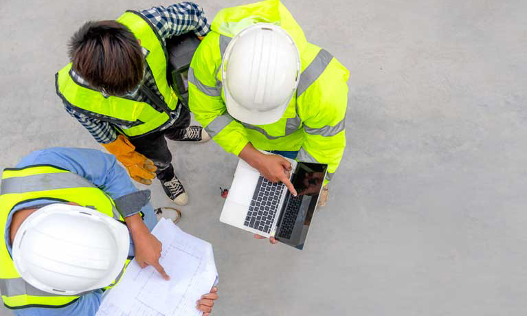 Three people wearing yellow vests and hard hats looking over construction plans.