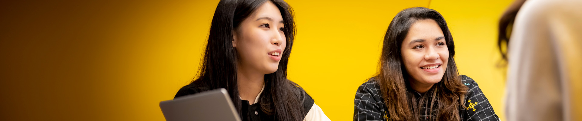 Two international students sat at a table with a laptop, against a painted yellow wall.