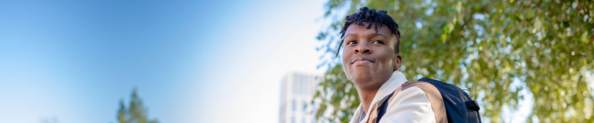 A student leaning against a wall with their phone in their hand, with trees and a blue sky in the background.
