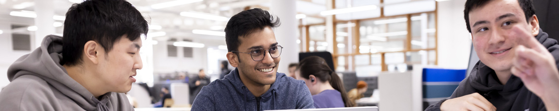 Three students smiling and working on laptops in an open study area.