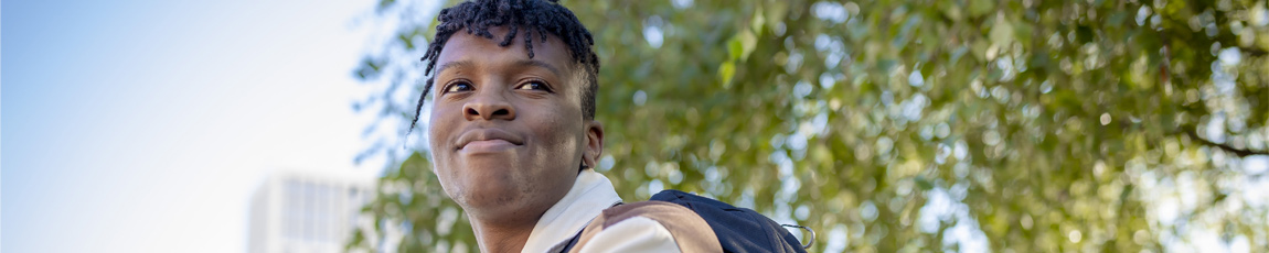 A smiling student with a blue sky and green trees behind them.