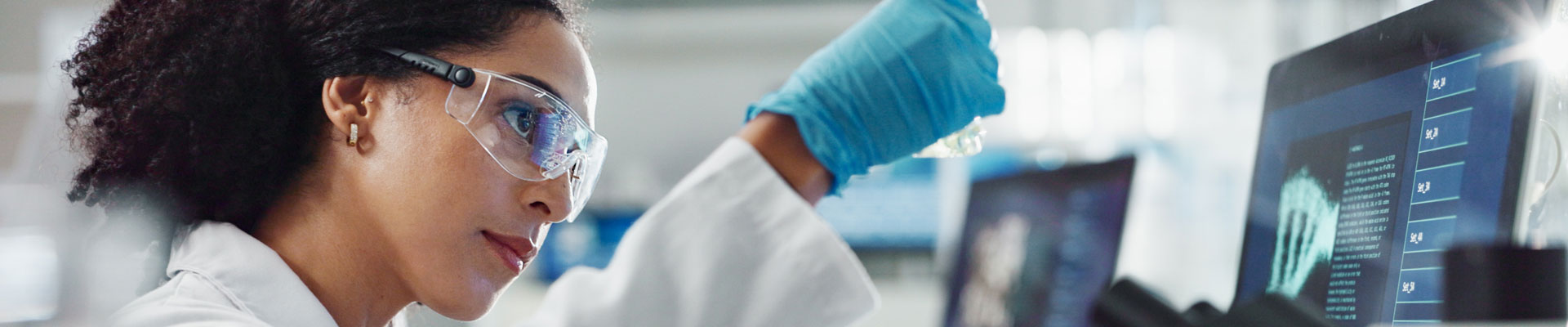 women doing lab work wearing white coat, goggles, and gloves