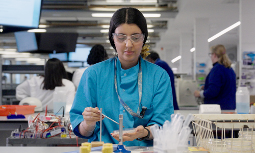 A student in a lab using a pipette and petri dish at a workstation.
