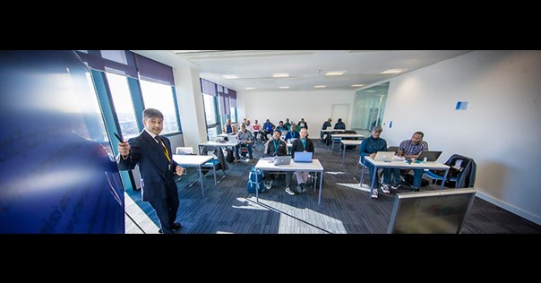 lecturer standing at the front of a classroom pointing at a screen and speaking to students