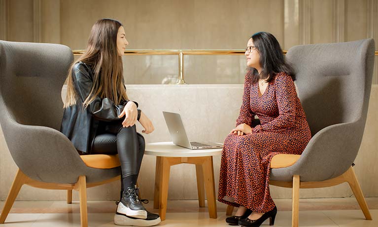 Two females sat down inside talking with an open laptop in between them