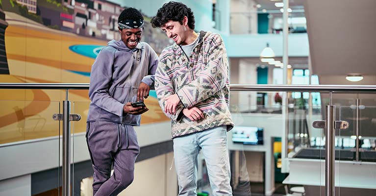 Two students leaning against a glass balcony looking down at a phone