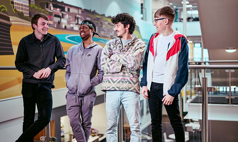 Four male students chatting whilst stood against a balcony inside a building