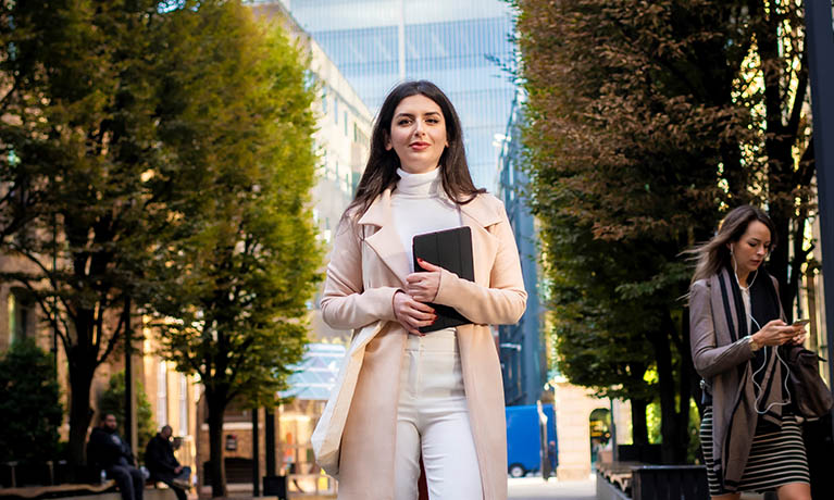 Woman holding notebook walking in front of trees.