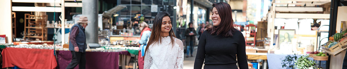 two females walking through an outdoor market with market stalls behind