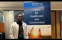 Ebenezer Akore Yeboah wearing a white jacket stood next to a banner for Coventry University's Research Centre for Healthecare and Communities