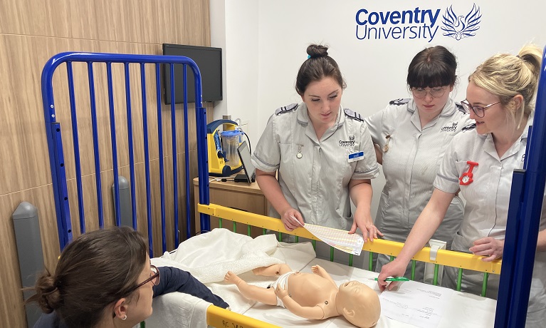 Three Coventry University students in nurses uniforms next to a cot containing a model of a baby