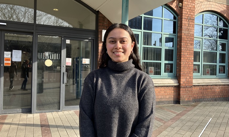 Nikita Lal wearing a black jumper smiling at the camera while stood outside Coventry University's William Morris building