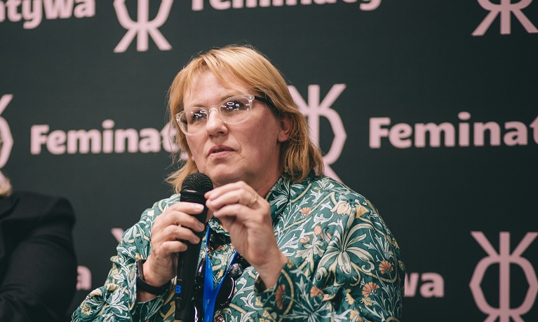 Coventry University assistant professor Alison Halford speaking into a microphone with the Feminatywa festival background behind her