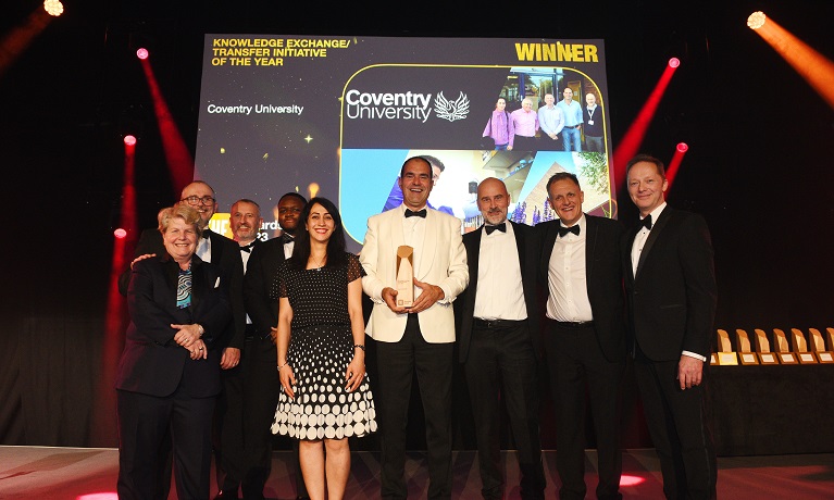 Sebastian Farnaud dressed in a suit holding the Times Higher Education award trophy on a stage alongside colleagues and hosts